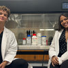 Two students in white lab coats sit on either side of a biosafety cabinet in a lab. Inside the cabinet, pipettes hang above lab supply containers, including bottles hand-labeled “PelliTect.”