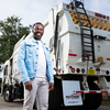 A smiling man wearing a light blue denim jacket and white pants stands in front of the rear of a large white garbage truck outdoors.